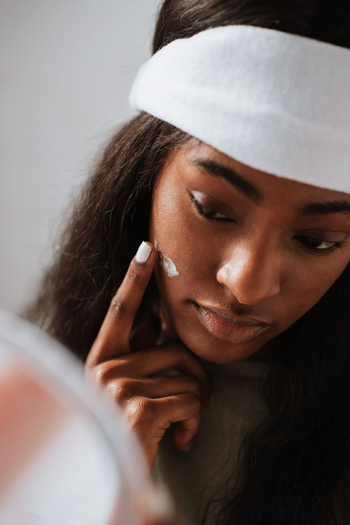 services-bg Crop young black female in headband applying hydrating cream on face against mirror on light background