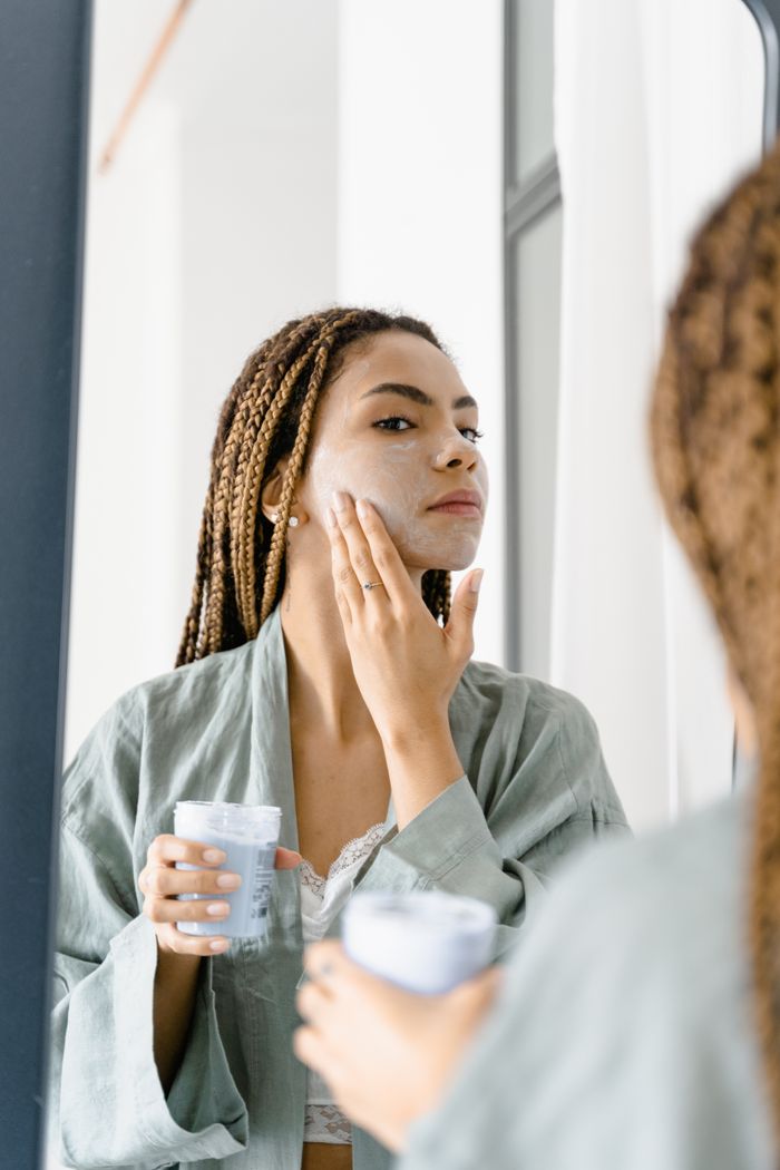our-story Woman in a green robe applying facial cream in front of the mirror for skin care.